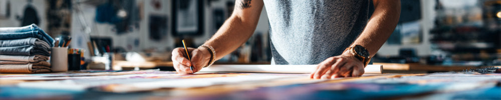 Person sketching on a large sheet of paper in a creative studio, surrounded by art materials and fabrics.